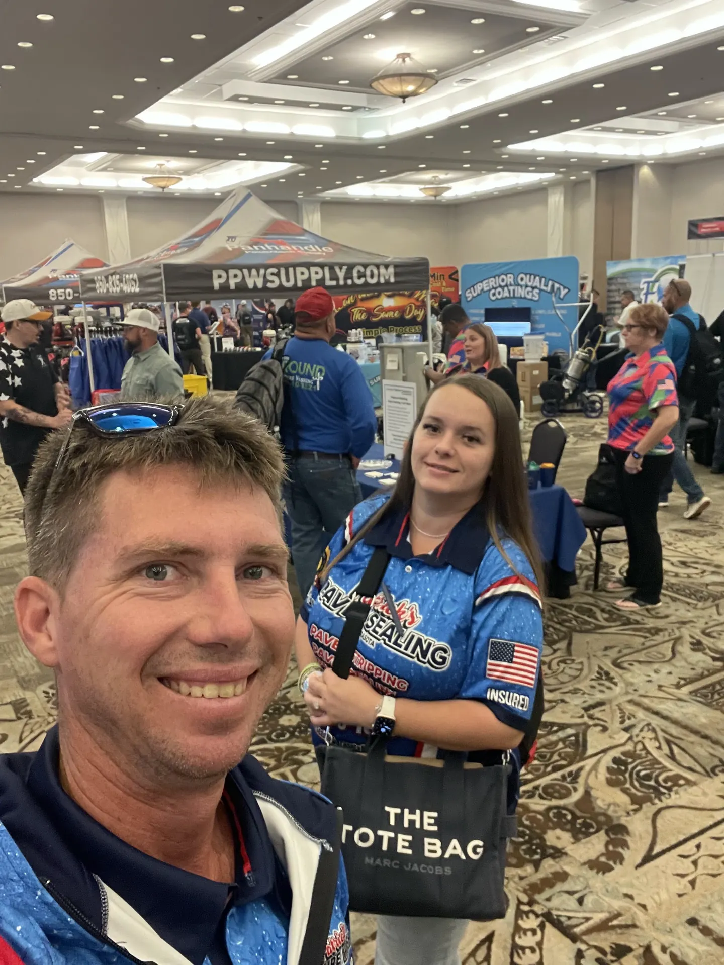 A smiling man taking a selfie in a trade show hall, in a blue branded jersey, with a woman in a matching jersey behind him