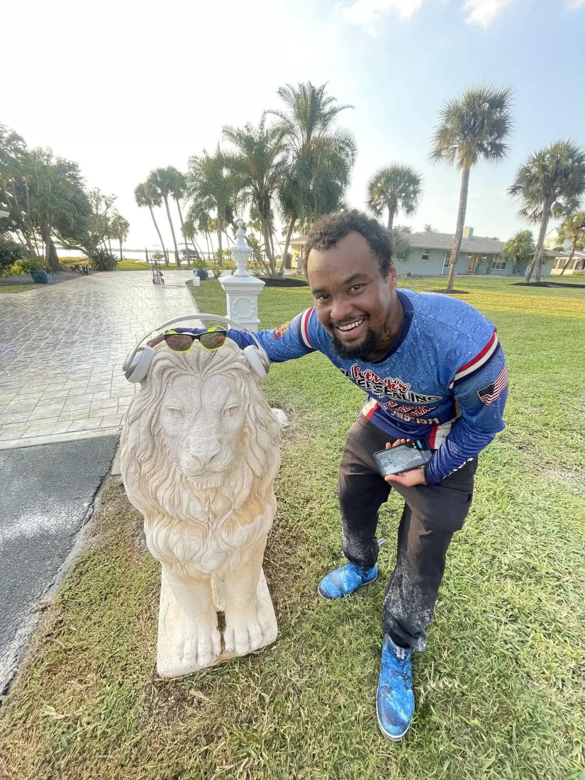 A smiling crew member in a blue branded jersey and shoe covers, leaning on a white lion statue on a lawn