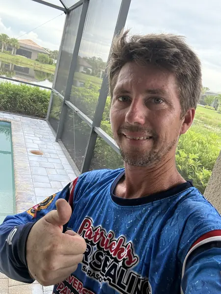 Smiling team member giving a thumbs up in a pool screen enclosure, wearing a blue branded team shirt