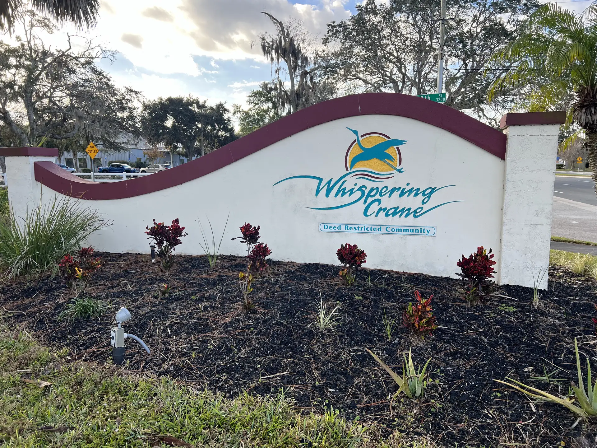 Curved white community entrance wall with a teal bird logo sign above mulch and red foliage plants