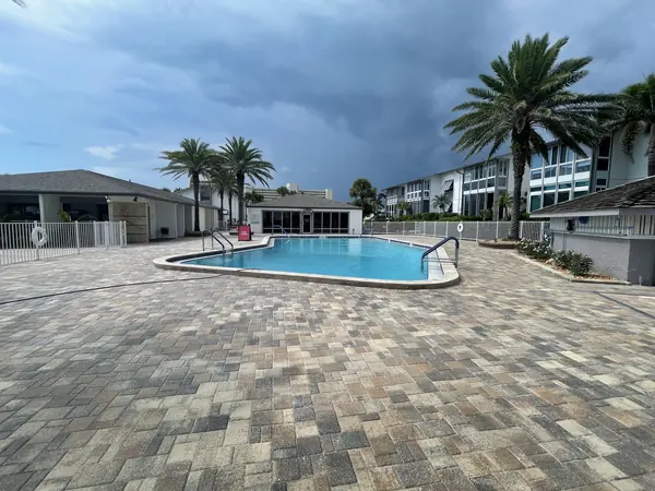 Clean brick paver pool deck with a blue pool, palm trees and condo buildings under a stormy sky