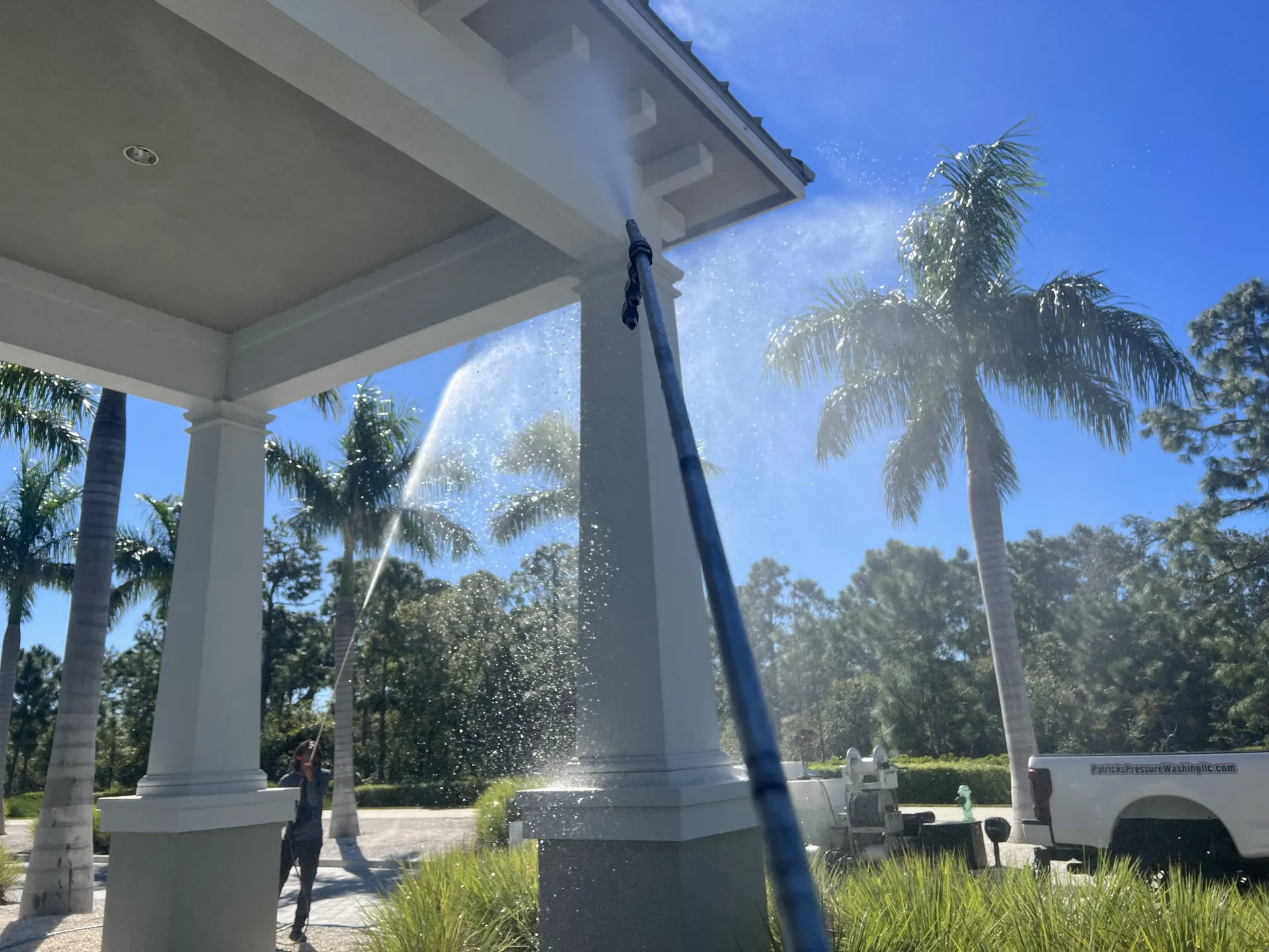 Worker in the distance using a long wand to soft wash the columns and underside of a covered entry canopy, spray lit by sun