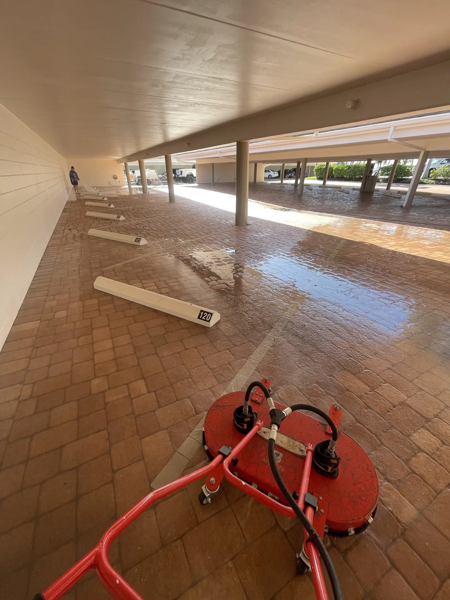 Red surface cleaner on wet brick pavers in a covered carport with numbered parking bumpers and a worker far behind