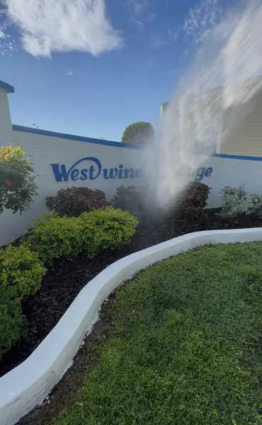 Water spraying across a community entrance wall and curved planter bed with a blue sign lettering partly visible