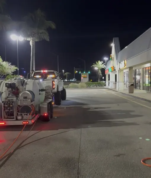 Night plaza with a pressure washing trailer and truck parked on wet asphalt, hose reels and tanks visible