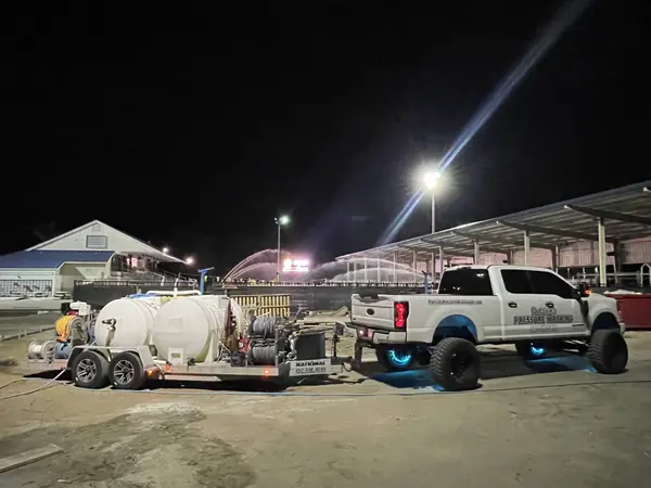 Night scene with a pressure washing truck and trailer on dirt ground beside a covered arena with floodlights and sprinklers