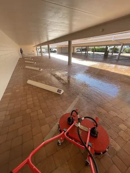 Red surface cleaner on wet brick pavers in a covered carport with numbered parking bumpers and a worker far behind