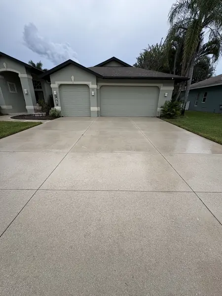 Clean light concrete driveway leading to a three car garage on a gray stucco home under a cloudy sky