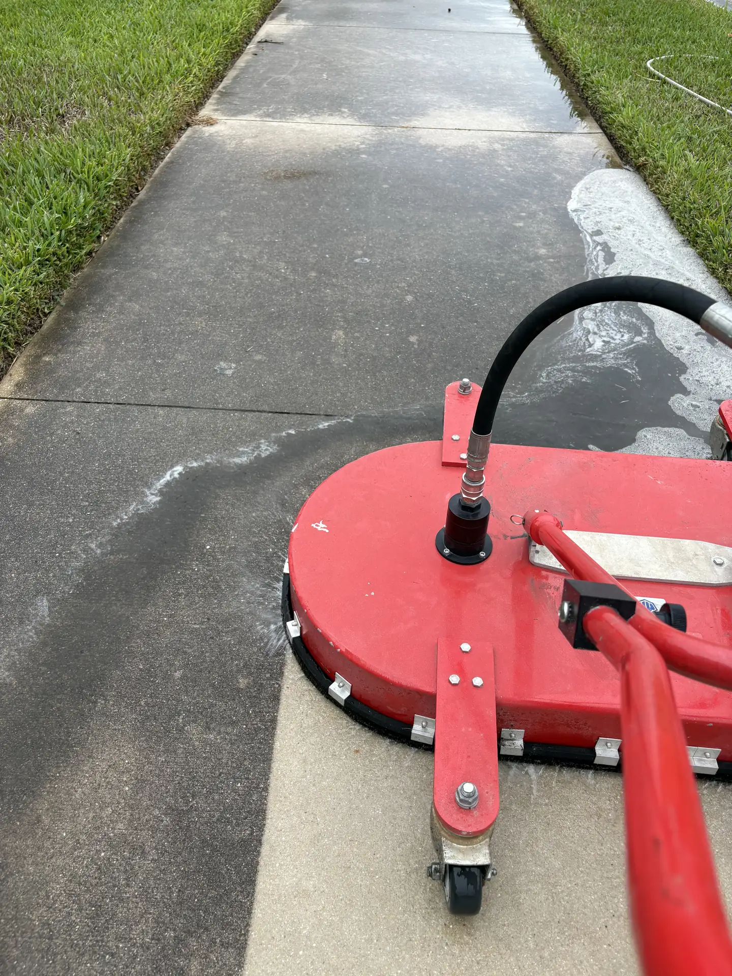 Red surface cleaner on a gray concrete walkway beside grass, leaving a lighter cleaned path