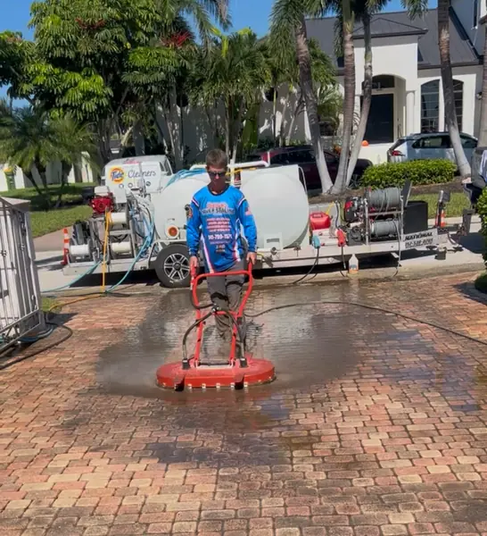 Worker pushing a red surface cleaner across a wet paver driveway with the pressure washing trailer behind him