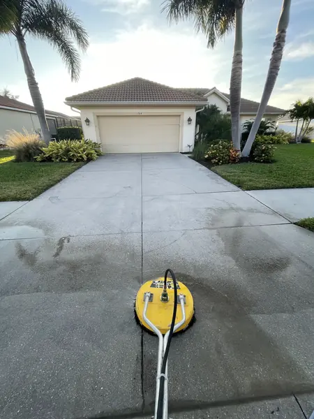 Yellow surface cleaner on a concrete driveway in front of a tan garage, half the driveway already washed