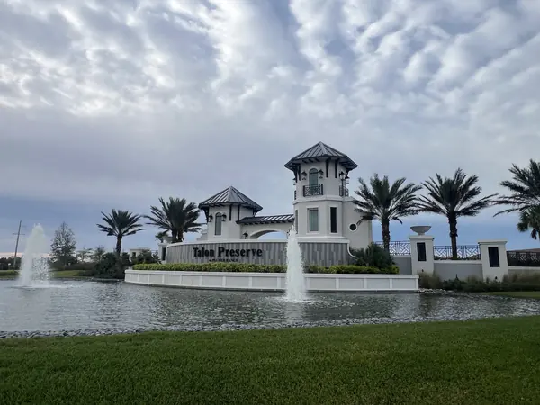 Community entrance sign and fountains beside a pond under a cloud streaked sky