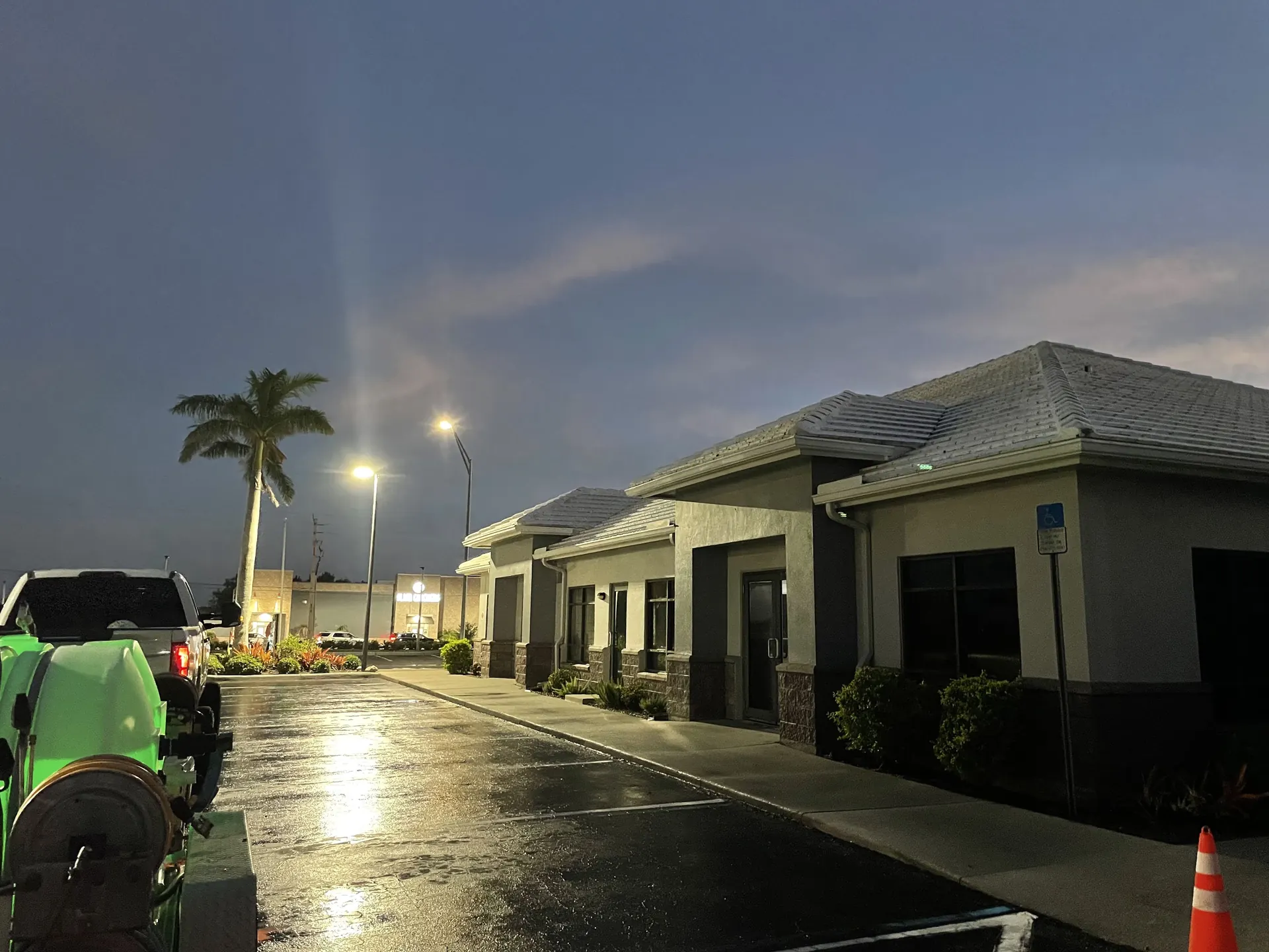 Wet asphalt parking lot at dusk in front of a row of tan commercial buildings with a green machine and cone at the left