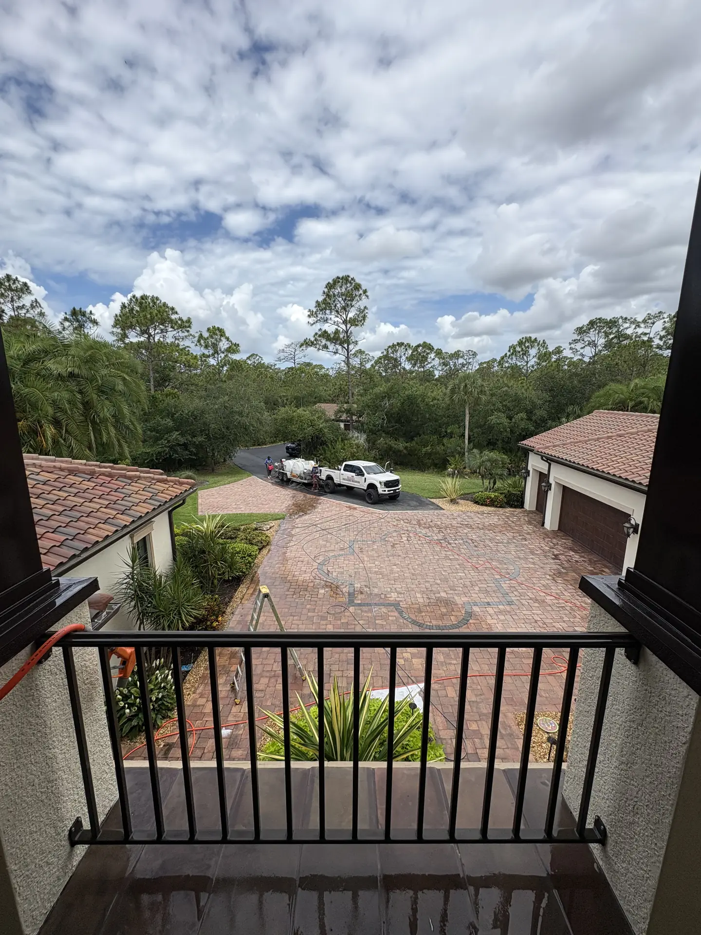 View from an upper balcony down to a paver driveway with ladders, hoses, and a pressure washing truck on the road