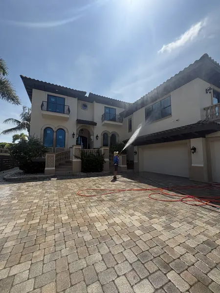 Worker spraying the upper walls of a large two story stucco home from a paver driveway with an orange hose