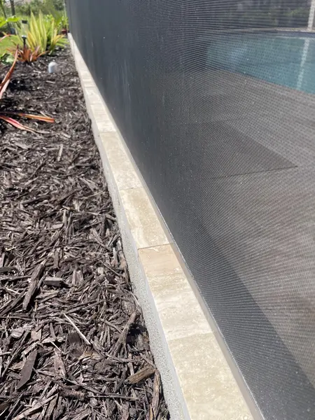 Pale dusty stone coping strip between a mulch bed and a dark pool screen, before view