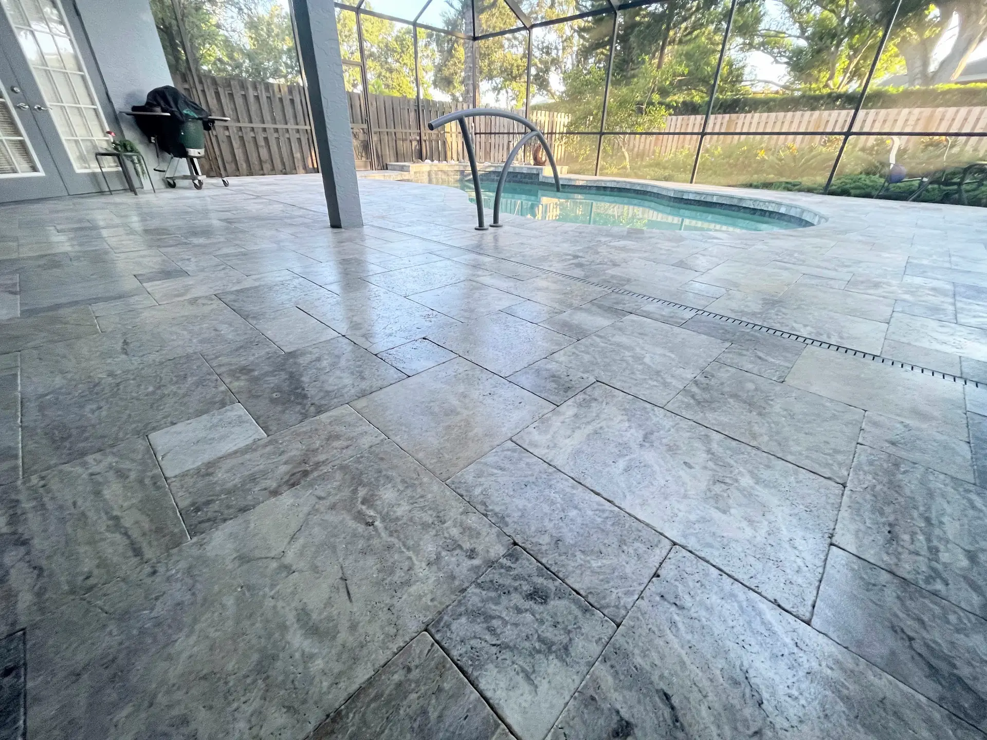 Silver grey travertine lanai floor in a pool screen enclosure with a grill, pool handrail and wooden fence visible