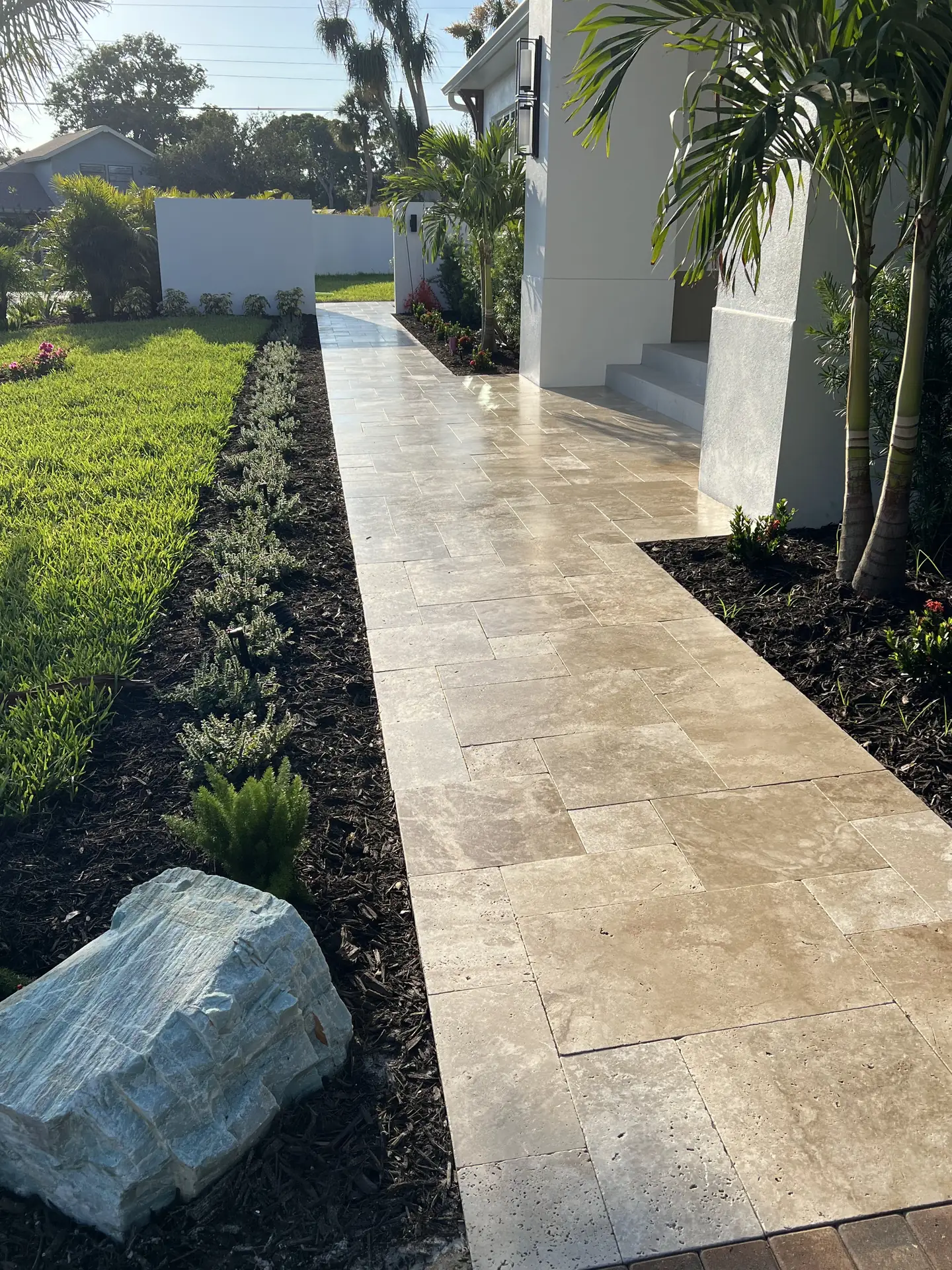 Travertine walkway to a home entrance between a lawn and dark mulch beds, with a pale boulder in the foreground