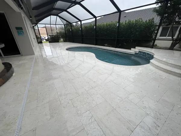 Cream stone pool deck inside a screen enclosure, wet from cleaning, with a pool, spa step and drain channel