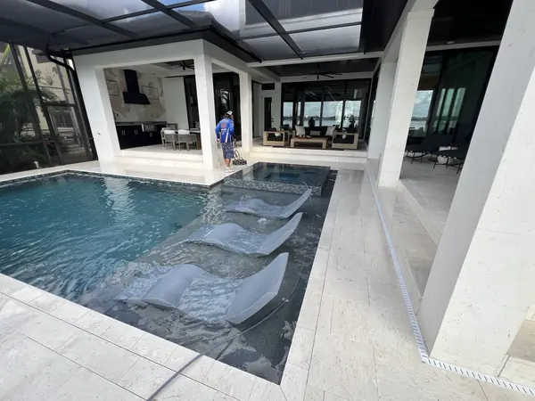 Worker guiding a surface cleaner beside a spa on a pale stone pool deck, covered lanai seating behind