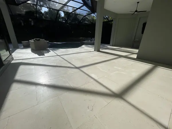 Pale stone floor in a screen enclosure with strong shadows from the cage frame, ceiling fan and pool in the distance