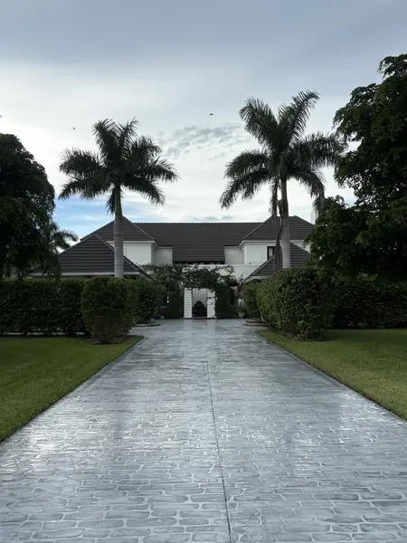 Wet, patterned gray driveway leading between two palm trees to a large house under an overcast sky