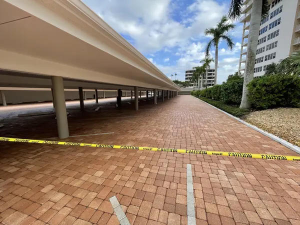 Long red brick paver drive beneath a multi-level covered parking structure beside a high-rise, with caution tape across it