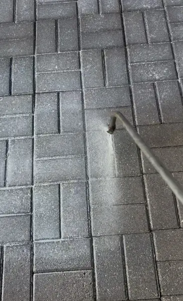 Pressure washer wand cleaning gray herringbone pavers, leaving a pale cleaned streak and white residue in the joints