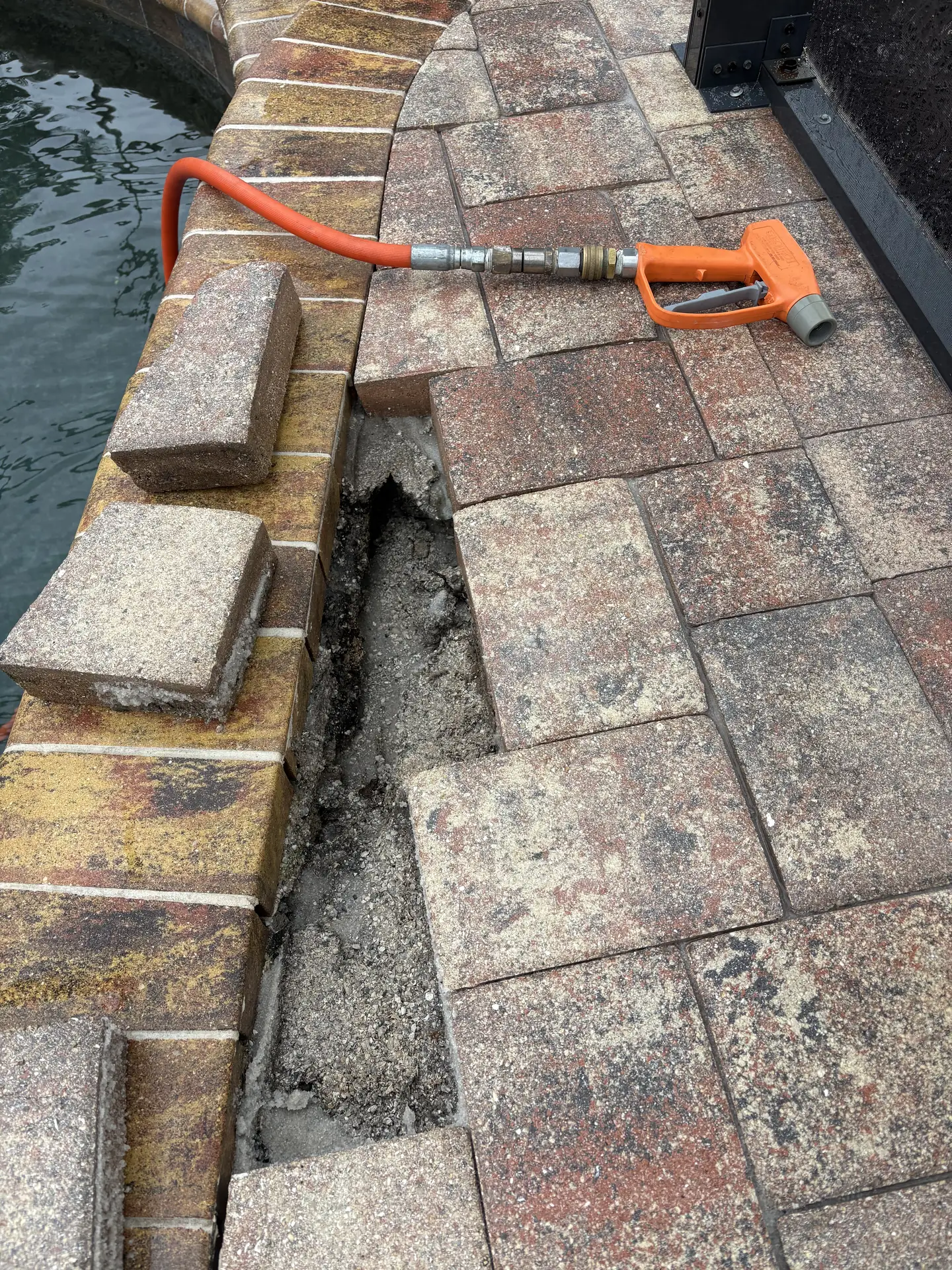 Pool edge pavers with a sunken gap along the coping and an orange pressure washer trigger gun lying on the deck