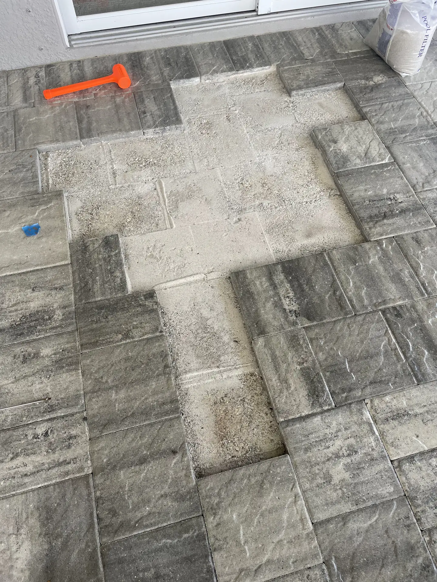 Gray pavers lifted beside a sliding door, bare bedding sand showing, with an orange mallet and a sand bag nearby