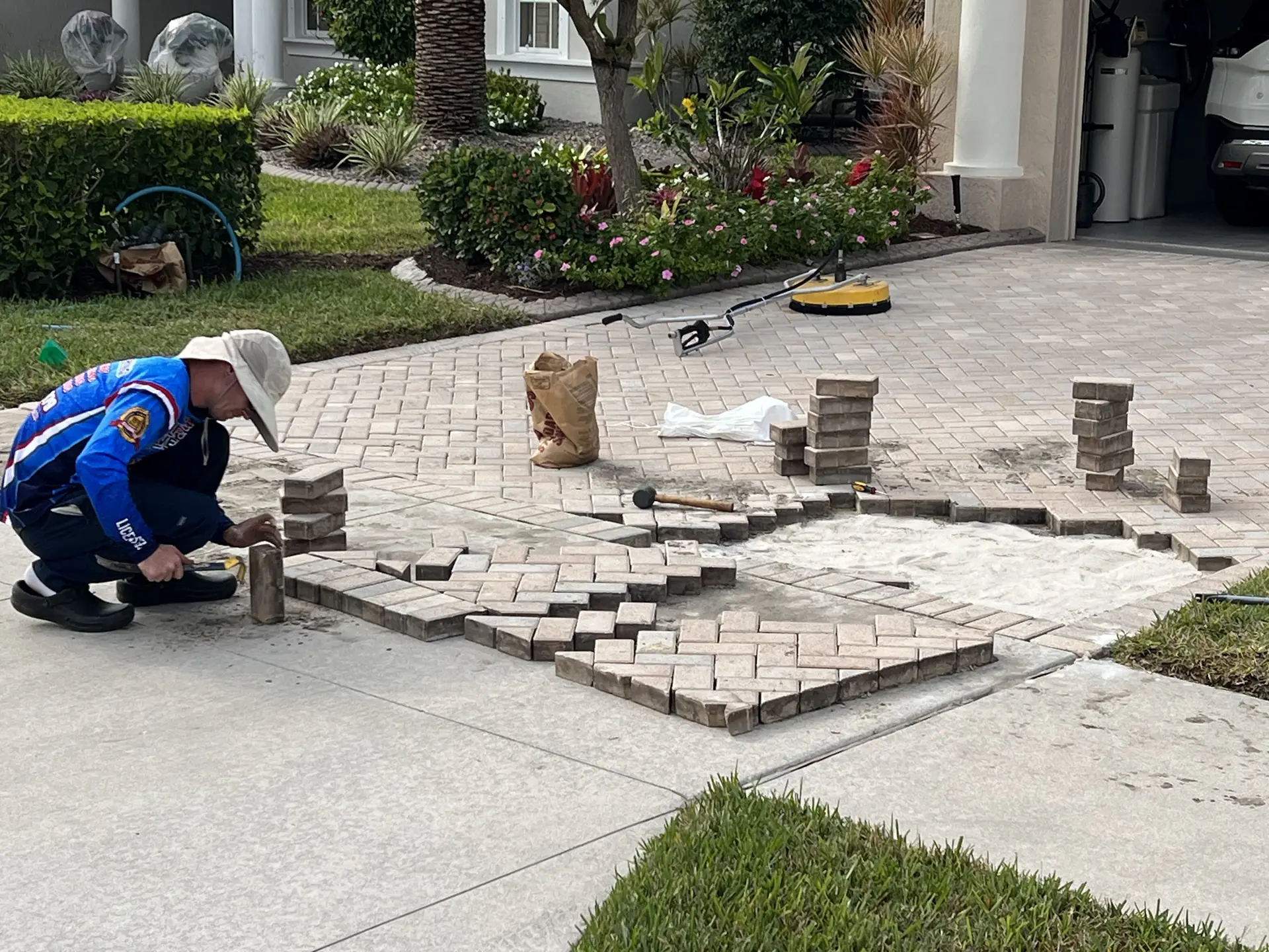 Worker in a blue shirt and sun hat kneeling to re-lay driveway pavers, with stacked pavers and a yellow cleaner nearby