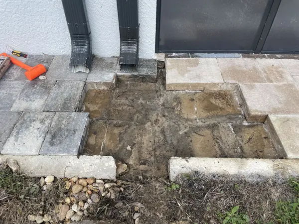 Section of pavers removed beside two downspouts, with bare earth base showing and an orange mallet on the surface