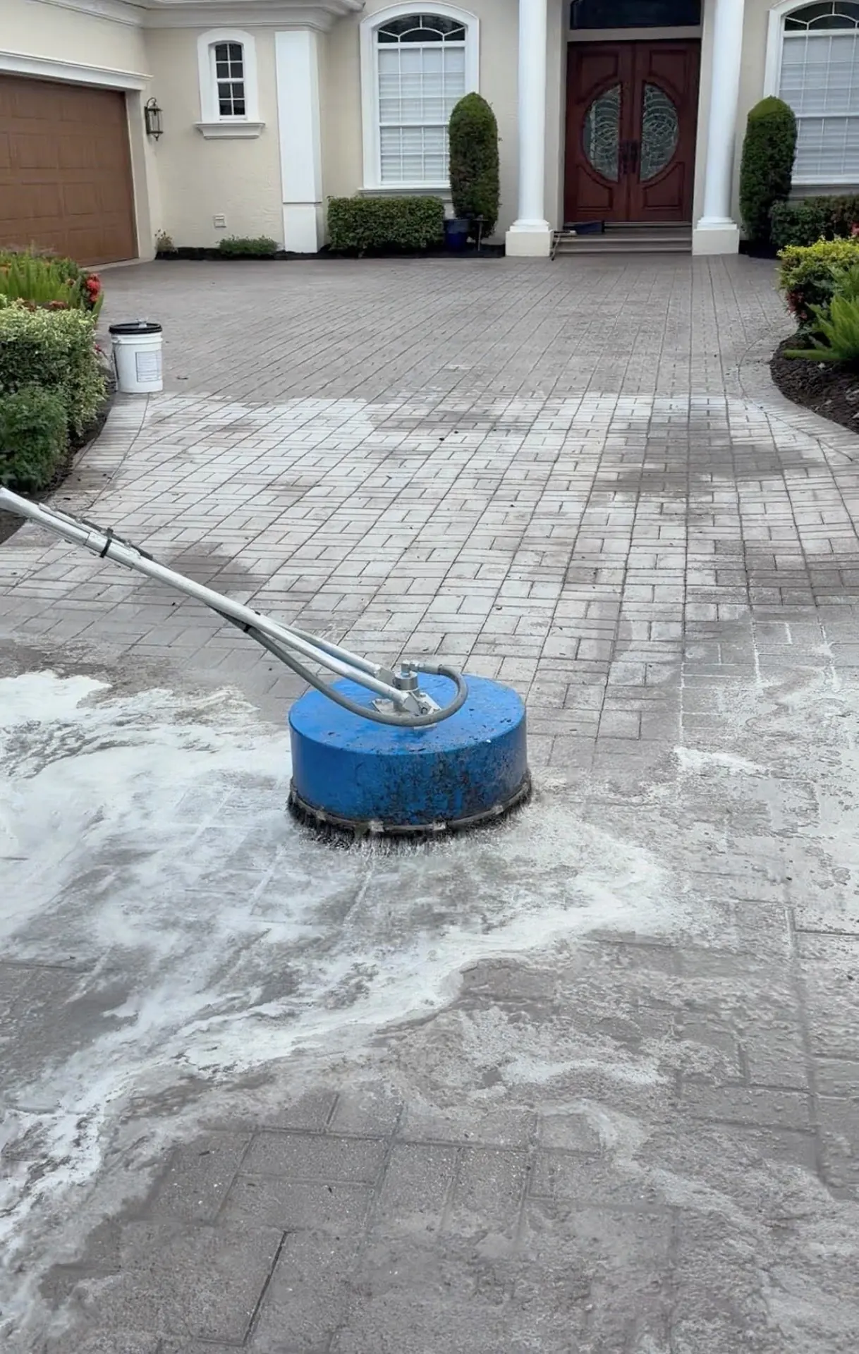 Blue surface cleaner on a large paver driveway with white foam, in front of a house with red double doors and a garage