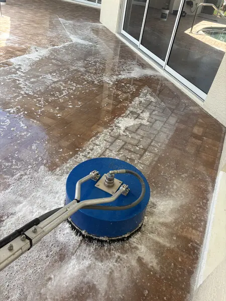 Blue rotary surface cleaner scrubbing wet brick pavers beside sliding glass doors, with white foam spreading around it