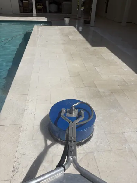 Blue rotary surface cleaner on a cream stone tile pool deck next to a pool, with a bucket and squeegee in the background