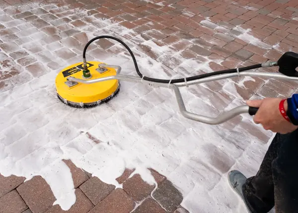 Yellow rotary surface cleaner on brick pavers coated in white foam, with the operator arm and legs partly in frame