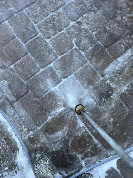Pressure wand spraying gray cobble-style pavers with foam, with an operator boot visible at the bottom