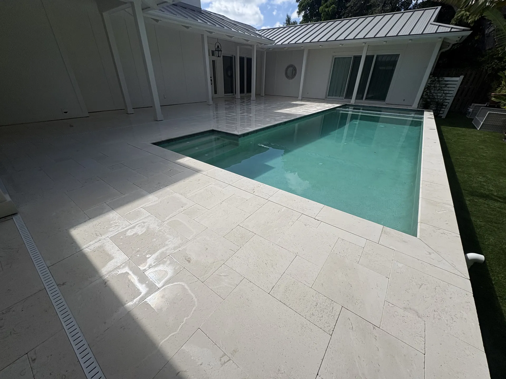 Pale smooth stone pool deck and lap pool beside a house with a metal roof, drain channel along the edge