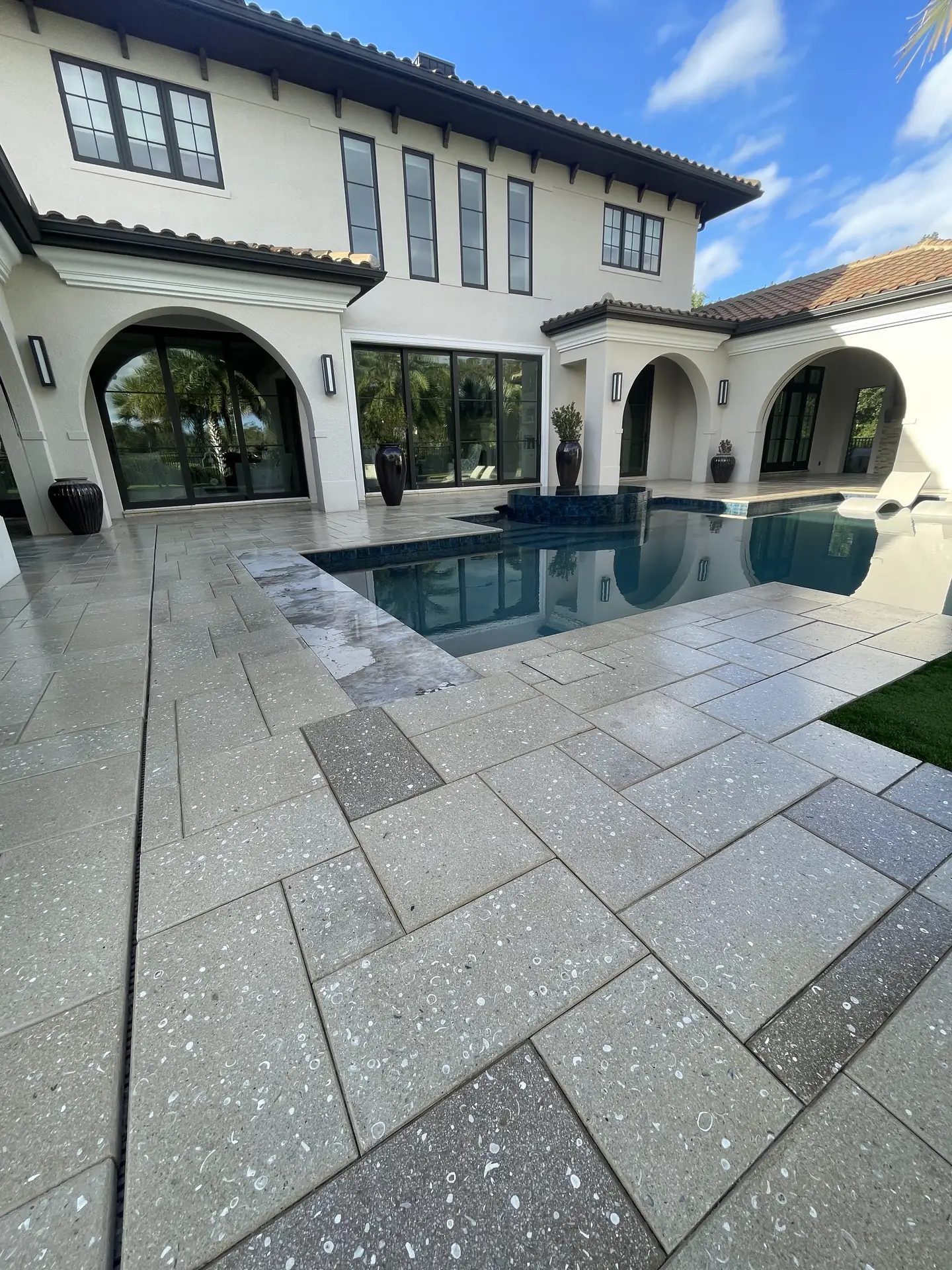 Large grey concrete paver pool deck with shell flecks in front of a Mediterranean style home with arches