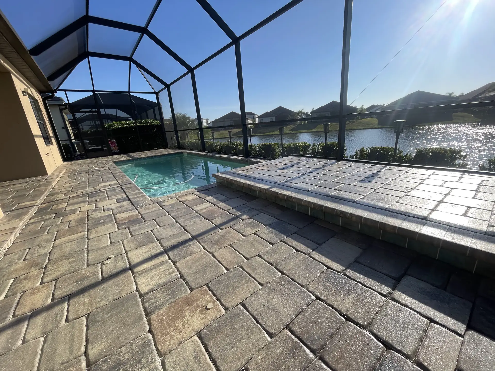 Brown tumbled paver pool deck in a screen enclosure beside a lake in low sun