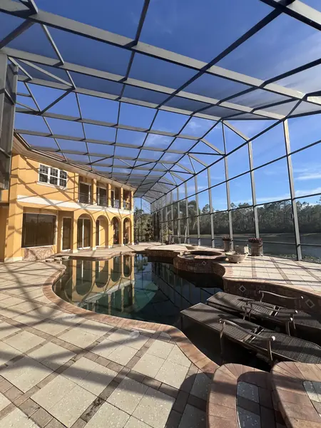 Inside a very large screen enclosure, a paver deck, pool and spa in front of a yellow home with a lake view