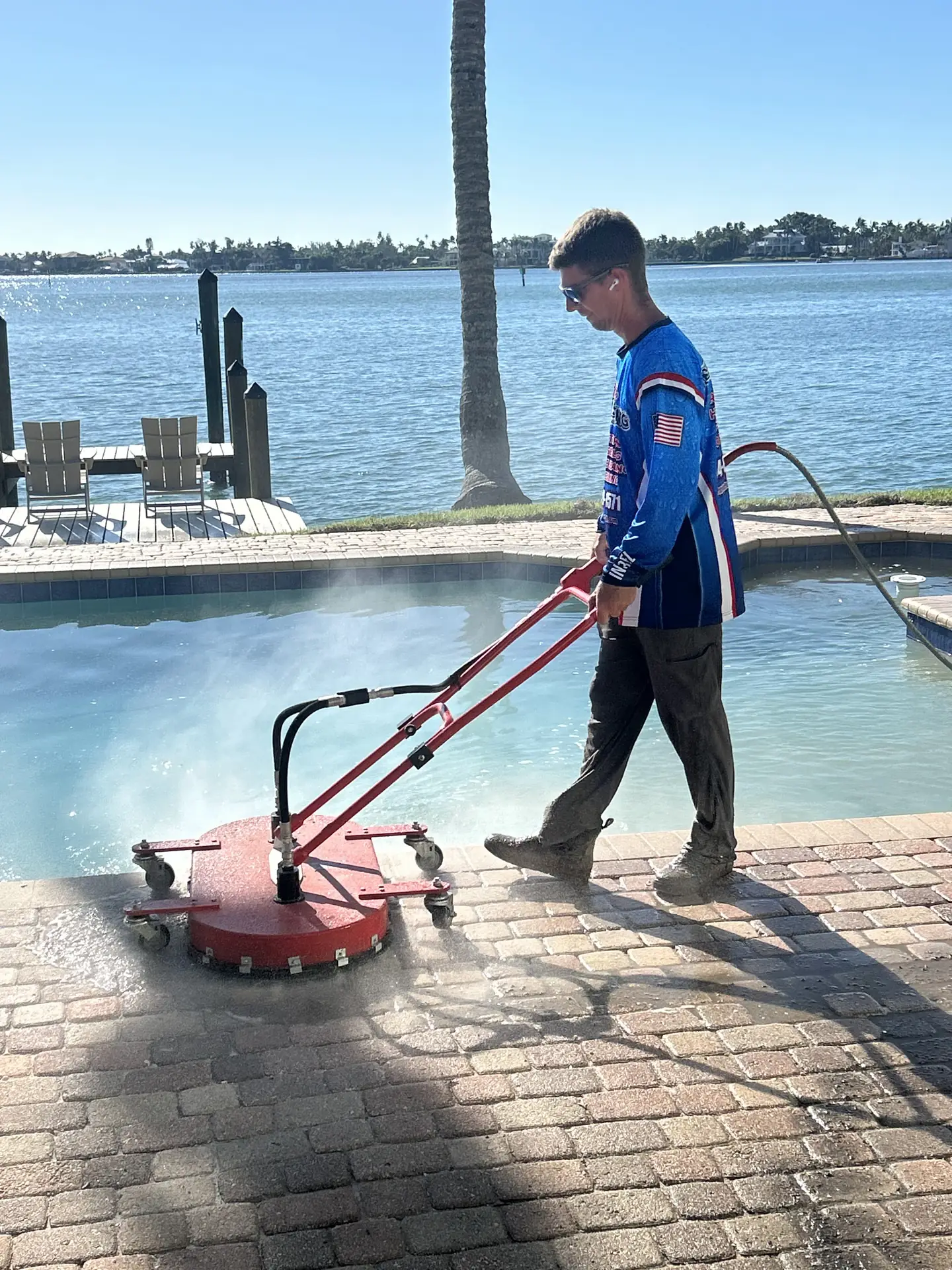 Worker in a blue team shirt walking behind a red surface cleaner on a brick paver pool deck beside the water