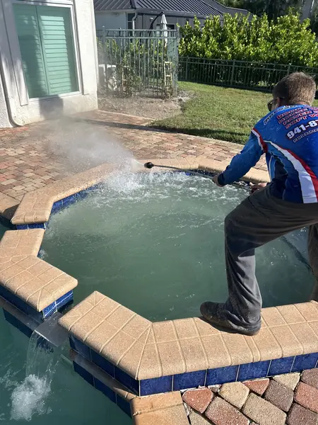 Worker in a branded shirt spraying a wand across the spa edge of a paver pool deck