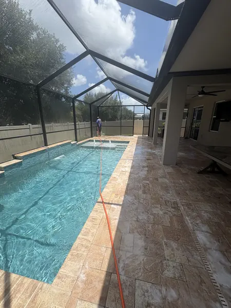 Narrow lap pool and travertine deck in a screen enclosure, a worker far away with an orange hose, foam on the stone